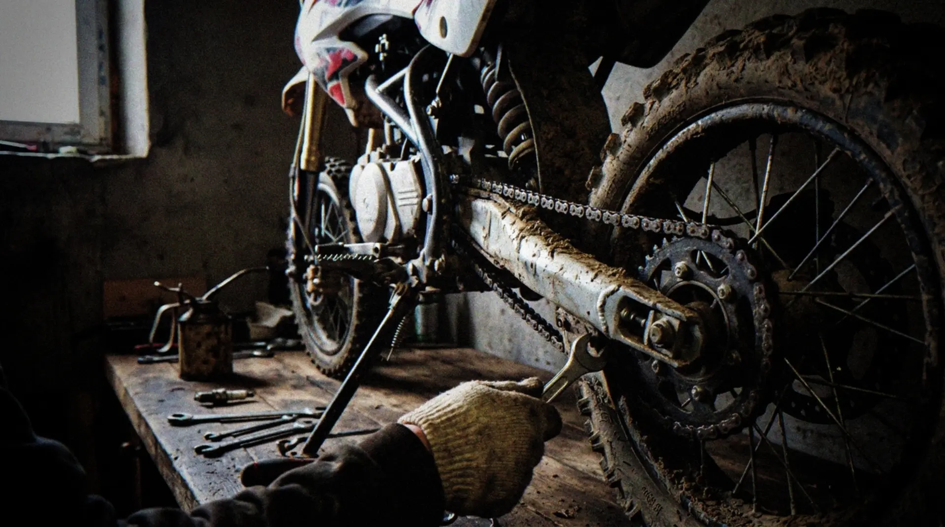 A close-up view of a dirt-covered pitbike in a garage workshop setting, with a mechanic's hand using a wrench to adjust the chain. Tools scattered nearby on a workbench, including an oil can, spark plug, and basic maintenance tools. The bike shows signs of recent use with mud splatter on tires. Natural indoor lighting from a small window creates realistic shadows across the maintenance area. The focus is on the mechanical components being serviced, capturing the everyday reality of pitbike maintenance.