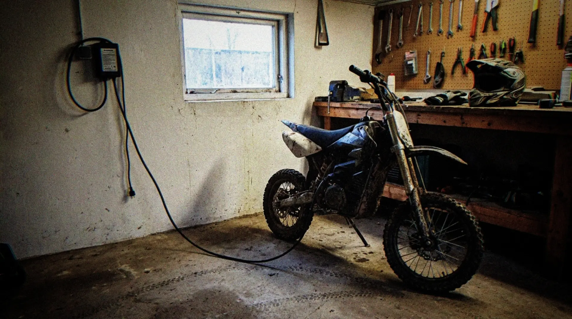 A documentary-style photograph of an electric dirt bike (pitbike) positioned next to a basic charging station in a home garage workshop. The bike is partially muddy from recent use, with visible electric cables connecting to a wall-mounted charger. Workshop tools hang on a pegboard in the background, with soft natural lighting coming through a small window. A riding helmet and gloves rest casually on a nearby workbench. The charging cable runs along the concrete floor, with tire tracks visible where the bike was ridden in. The scene captures the practical reality of maintaining and charging an electric pitbike in a typical home setting.