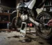 A small, dusty pit bike motorcycle propped on its stand in a home garage setting, with basic tools scattered on a workbench nearby. A mechanic's hands visible from the side as they check the chain tension. Natural indoor lighting shows specks of dust in the air. The workspace appears lived-in with some oil stains on the concrete floor, a few wrenches, screwdrivers and an oil container visible. The bike is slightly muddy from recent use, with dirt visible on the tires and undercarriage. A simple garage backdrop with typical household items on shelves in the soft-focus background.